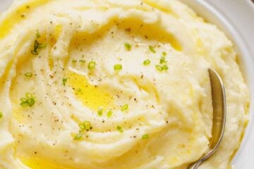 A bowl of sour cream mashed potatoes topped with melted butter and freshly chopped chives, with a spoon.