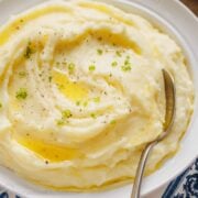 A bowl of sour cream mashed potatoes topped with melted butter and freshly chopped chives, with a spoon.