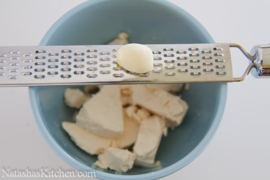 Garlic cloves being grated into blue bowl with cows cheese