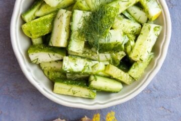A bowl of fresh cucumber salad in a bowl garnished with dill, dill and garlic cloves beside it