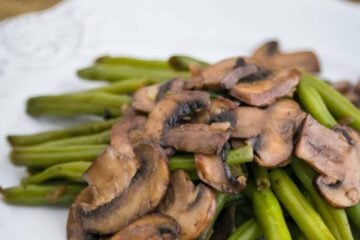 Green beans and mushrooms on a white plate with raw mushrooms behind the plate