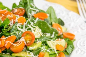 A plate of spinach salad with tomatoes and cucumbers in the background and a fork beside the plate