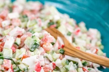A blue bowl of crab salad with cucumber and tomato with a wooden spoon in it