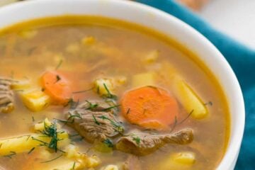 A white bowl of beef, barely and pickle soup with bread in the background