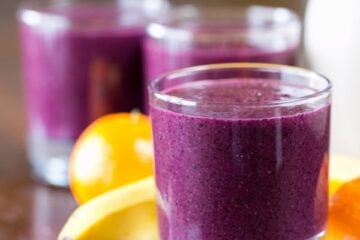 A close up of a glass cup with a blueberry smoothie with a banana and mandarines behind it