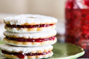 A close up of three jam-filled sandwich cookies on a plate with a jar of jam in the background