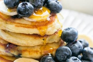 Stack of blueberry pancakes on a plate topped with syrup, blueberries, and sliced bananas.