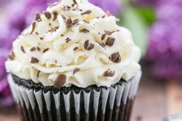 A close up of a dark chocolate cupcake with white chocolate frosting with flowers behind it