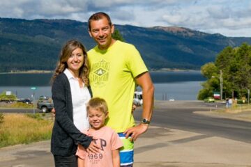 Natasha, her husband and son smiling at the camera with a body of water behind them