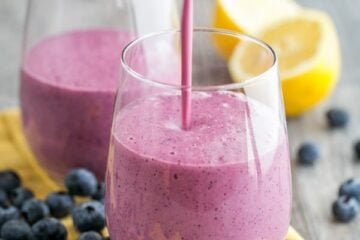 Two glass cups with a blueberry mango smoothie and blueberries and a halved lemon around the cups