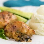 A plate of baked mushroom stuffed chicken thighs besides mashed potatoes and cucumbers