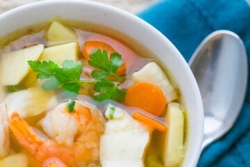 A white bowl of fish and shrimp soup, ukha, with a spoon, and bread bun besides it