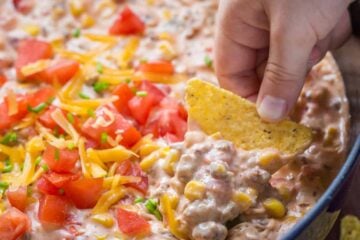 Sausage Queso Dip in pan topped with tomato, green onion, and cheese with a hand dipping a chip in it.