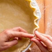 Hands crimping the edge of an unbaked homemade pie crust in a white pie dish