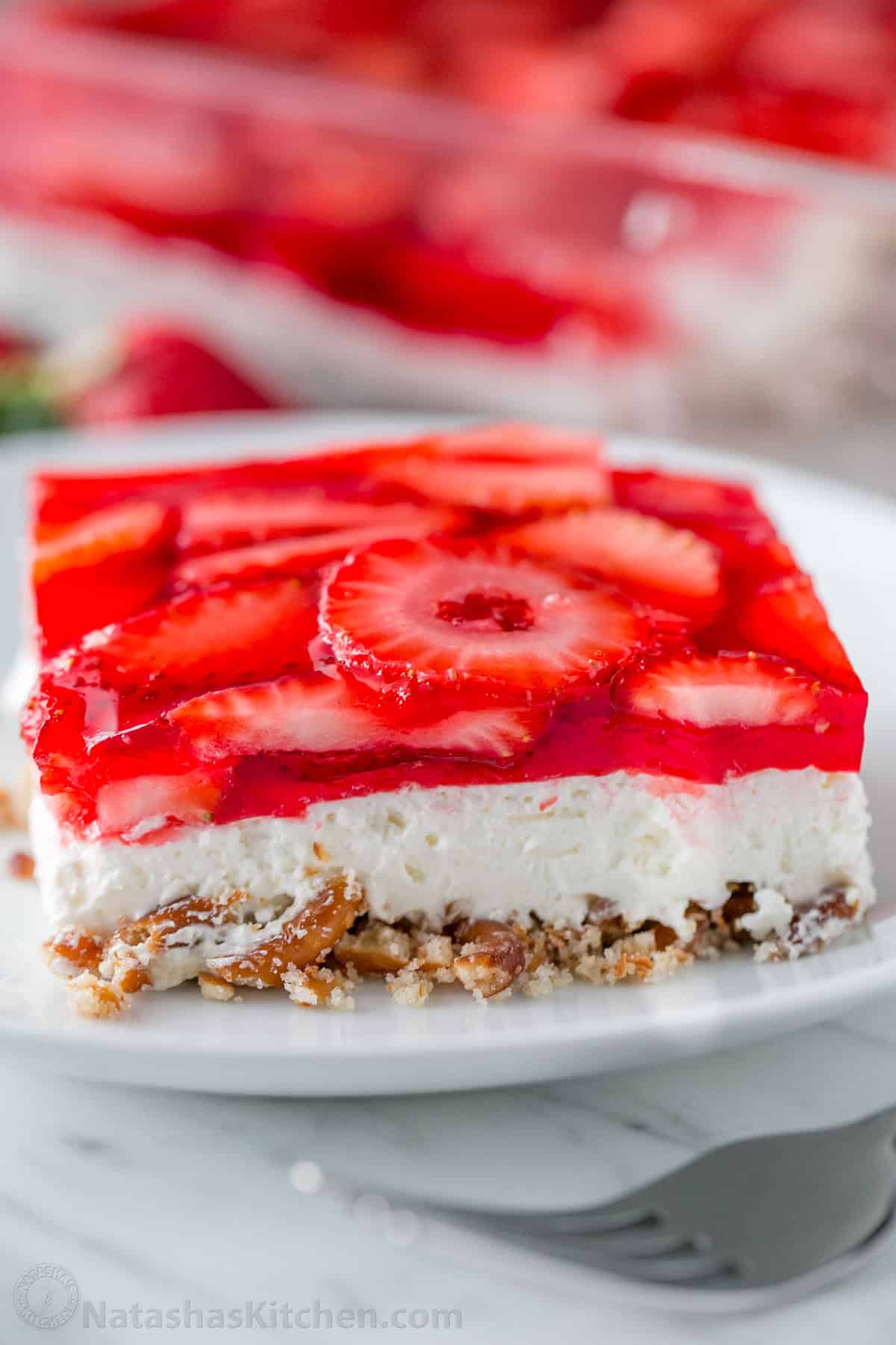 Easy Strawberry Pretzel Salad is a crowd-favorite dessert recipe at parties and picnics! Easy make-ahead directions everyone will love! A slice of Strawberry Pretzel Salad on a white plate with the whole dessert in the background