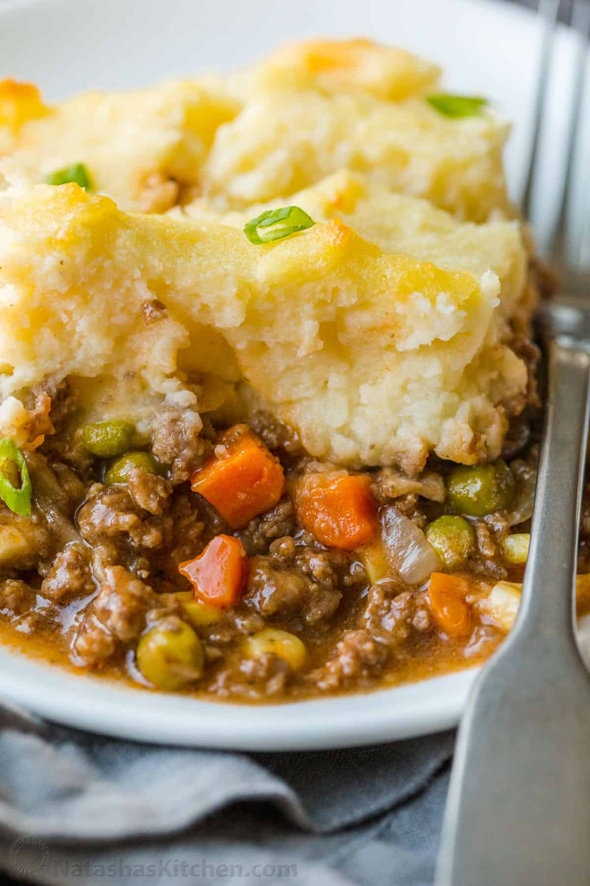 A homey and comforting Shepherd's Pie recipe with a meat and vegetable gravy sauce under a crown of creamy parmesan mashed potatoes. Slice of English Cottage casserole with ground beef, carrots, peas, and mashed potatoes