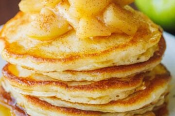 A stack of apple pancakes on a white plate, topped with sauteed cinnamon apples, with green apples in the background.
