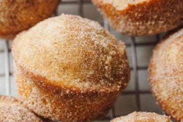Cinnamon muffins coated in cinnamon sugar on a wire rack, with one muffin on its side with a bite missing.