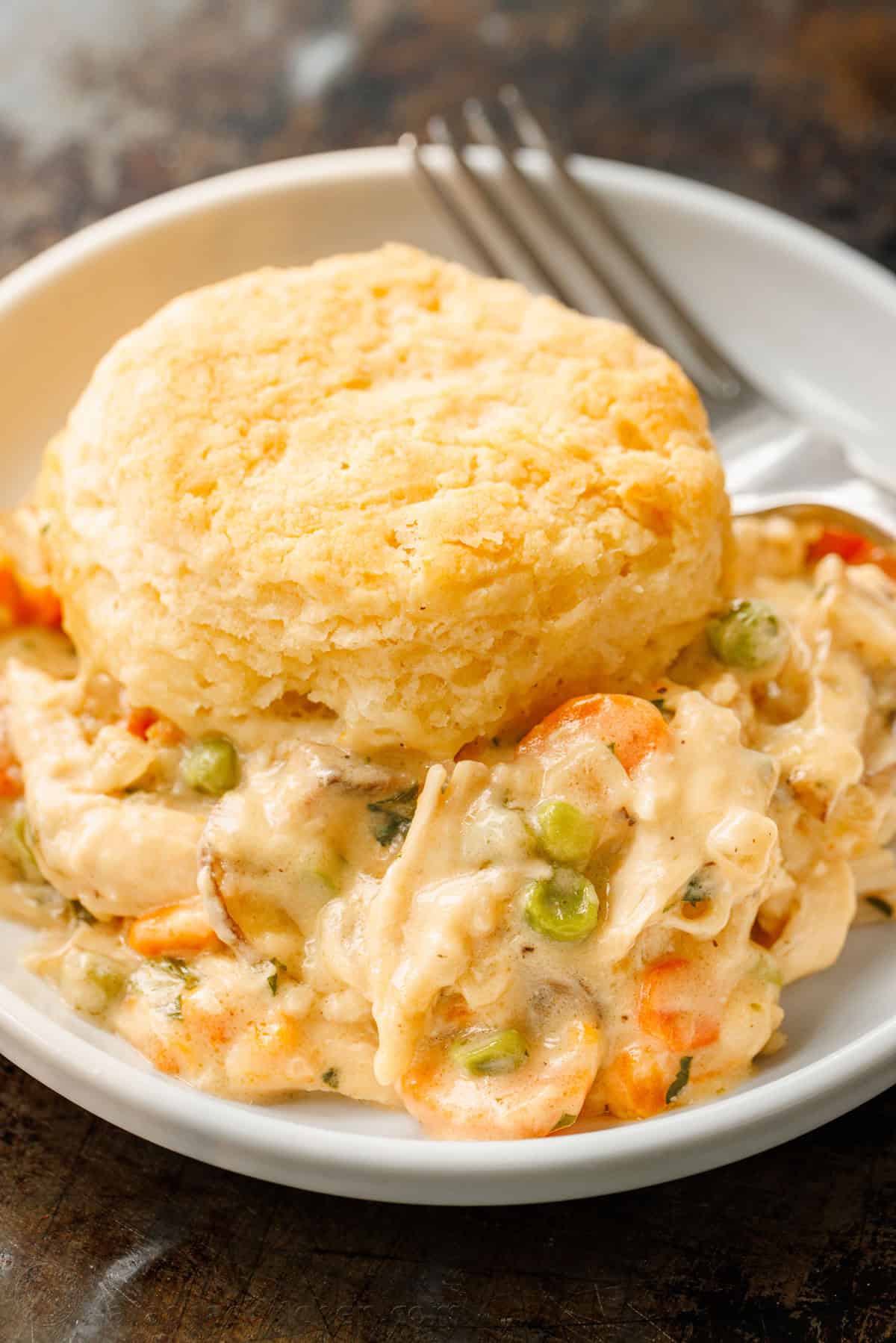 Chicken Pot Pie Casserole has all the same cozy flavors we love, but it's so easy to bake in a casserole dish, topped with flaky biscuits. A serving of chicken pot pie casserole topped with a biscuit in a bowl, next to a fork.