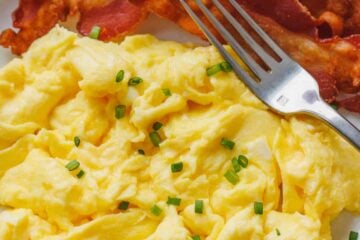 Fluffy scrambled eggs on a plate garnished with chives, next to crispy bacon and a fork.