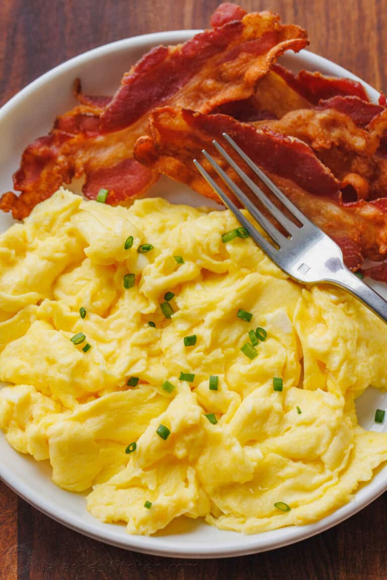 Fluffy scrambled eggs on a plate garnished with chives, next to crispy bacon and a fork.