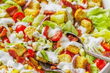 Close up of homemade garden salad in a large white bowl topped with croutons and drizzled with Ranch dressing, with two serving spoons.