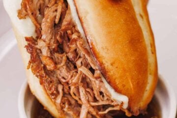A hand dipping a shredded beef sandwich into a ramekin of beef drippings.