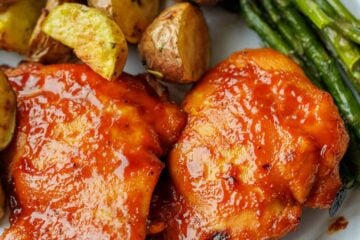 Two baked BBQ chicken thighs on a white plate next to roasted potatoes and asparagus.