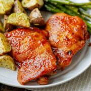 Two baked BBQ chicken thighs on a white plate next to roasted potatoes and asparagus.