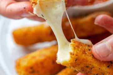 Two hands pulling apart a mozzarella stick with a plate of cheese sticks in the background.