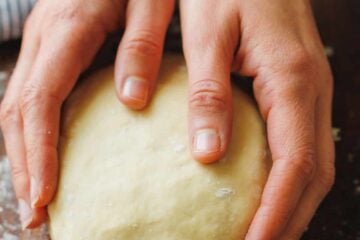 Two hands holding a ball of homemade pizza dough on a floured wooden countertop.