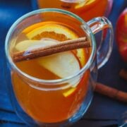 Apple cider garnished with apple and orange slices and a cinnamon stick. Served in a glass mug.