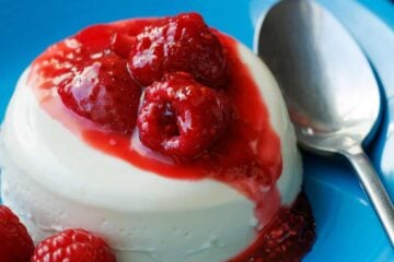 Panna Cotta with berry sauce drizzled on top, sitting on a blue plate with a spoon.
