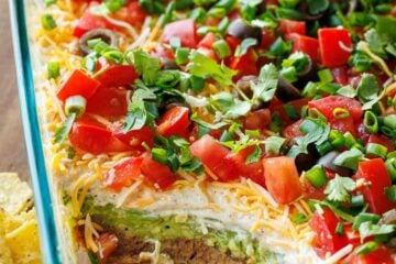 7-Layer Dip in a glass pan, showing each layer, refried beans, sour cream, cheese, tomatoes, green onion, guacamole, black olives with tortilla chips scattered around