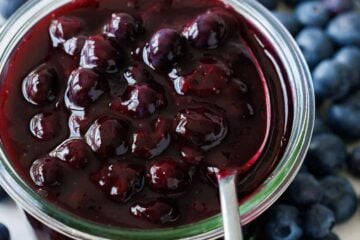 Homemade blueberry sauce in a glass jar with a spoon showcasing the whole blueberries. Fresh blueberries scattered on the counter around the jar.