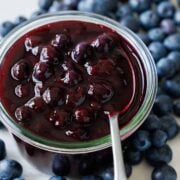 Homemade blueberry sauce in a glass jar with a spoon showcasing the whole blueberries. Fresh blueberries scattered on the counter around the jar.