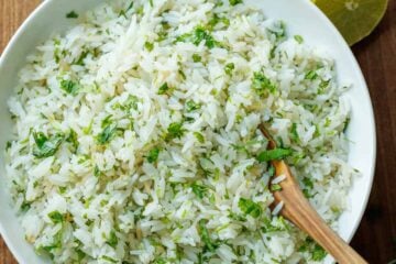 Cilantro lime rice in a large white serving bowl with a wooden spoon and slice limes