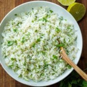 Cilantro lime rice in a large white serving bowl with a wooden spoon and slice limes