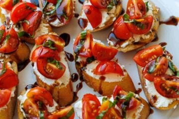 tomato goat cheese crostini on a white plate with cherry tomatoes and balsamic glaze on top