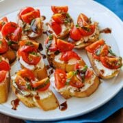 tomato goat cheese crostini on a white plate with tomatoes, basil, and balsamic glaze drizzle