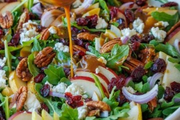 Pear Salad being dressed with Balsamic Vinaigrette in a white bowl, loaded with a variety of pears, red onion, goat cheese, cranberries, and toasted pecans