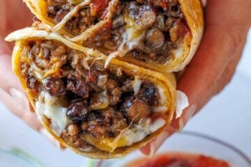 Beef burrito sliced in half to showcase the beef, melty Mexican cheese black beans, and Rotel tomatoes packed inside with bowls of salsa and guacamole toppings on the side.