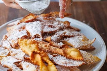 Dusting Angel Wing Cookies/ Polish Chrusciki with powdered sugar on a white platter