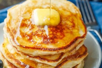 Sourdough Pancakes stacked on a plate with a pad of butter and maple syrup being poured over top.