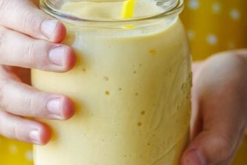 hands holding a yellow mango smoothie in a mason jar with yellow polka-dot background