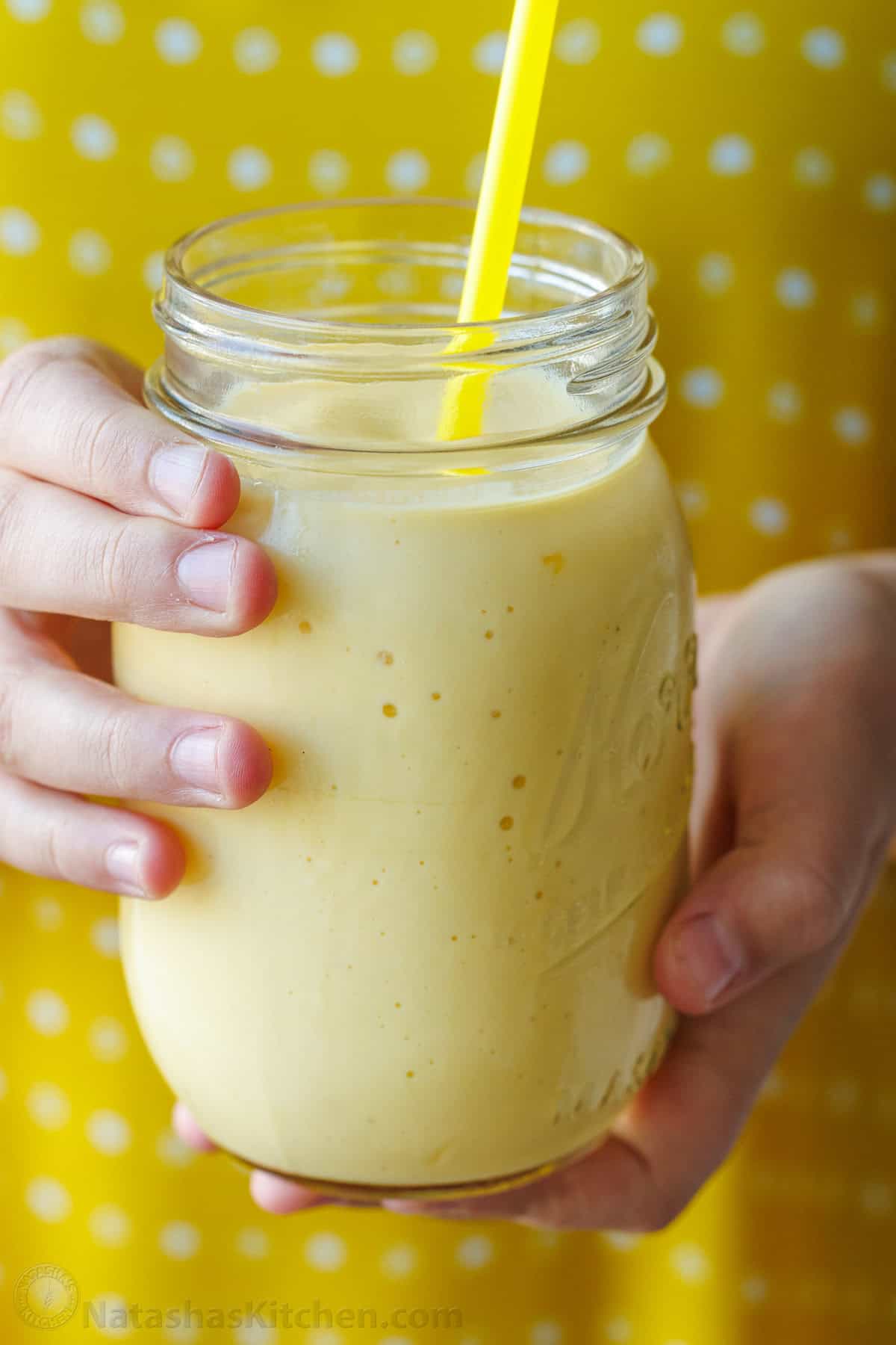 hands holding a yellow mango smoothie in a mason jar with yellow polka-dot background