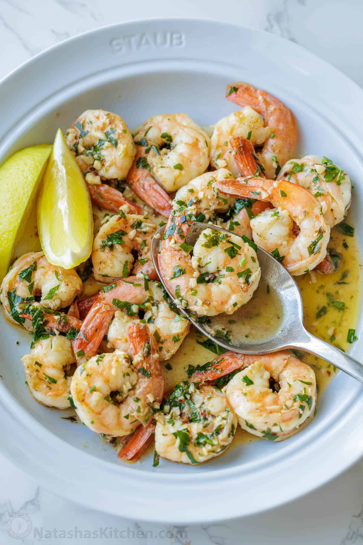 Spoon serving shrimp scampi from a white dish with garlic butter sauce and lemon wedges
