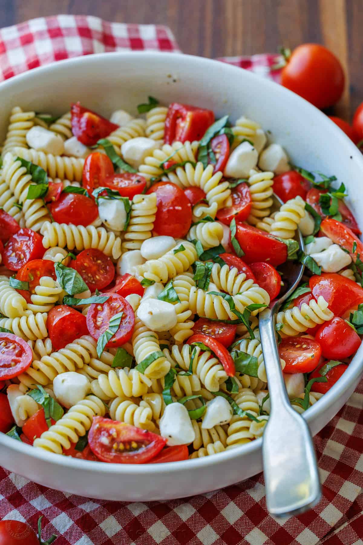 Caprese pasta salad combines the classic Caprese flavors of tomato, mozzarella and basil with Italian pasta salad. Perfect for BBQ potlucks. Caprese Pasta Salad on a red plaid napkin with cherry tomatoes around