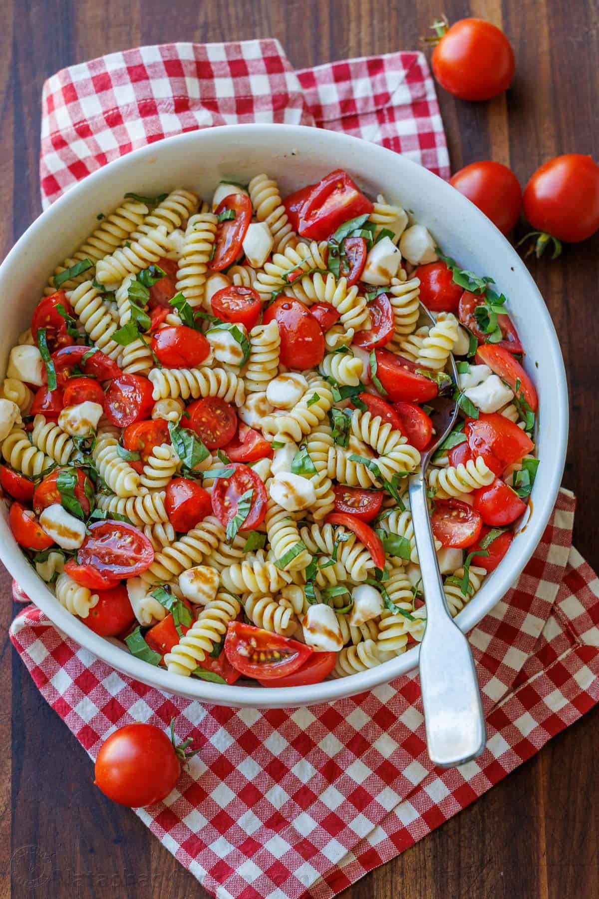 Caprese pasta salad combines the classic Caprese flavors of tomato, mozzarella and basil with Italian pasta salad. Perfect for BBQ potlucks. Caprese pasta salad recipe in a white bowl with a spoon for serving.
