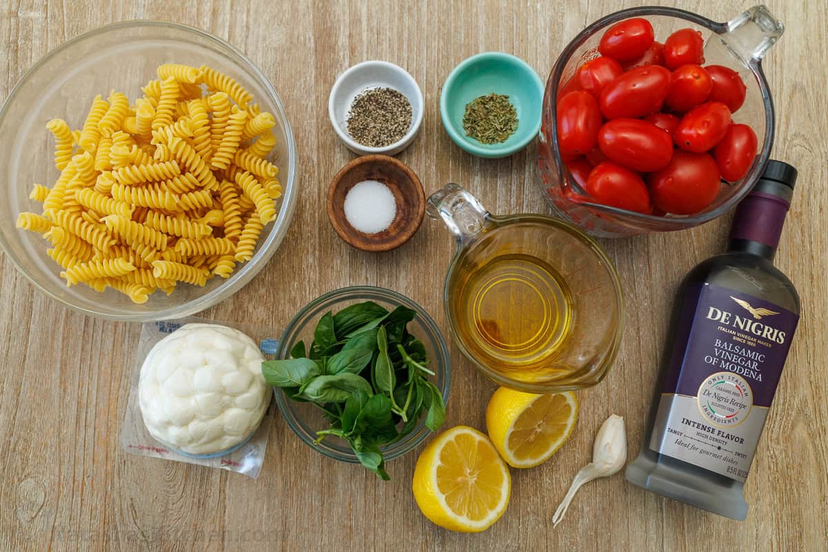 Caprese pasta salad combines the classic Caprese flavors of tomato, mozzarella and basil with Italian pasta salad. Perfect for BBQ potlucks. ingredients for Italian side dish recipe including fusilli, mozzarella balls, cherry tomatoes, basil, balsamic glaze, lemon juice, olive oil, salt, pepper, oregano, and garlic cloves